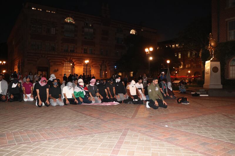 A group of over a dozen students kneel and bow, organized in rows while a single student kneels alone in front. They are in an open plaza space, facing west for prayer, as is tradition in Islam