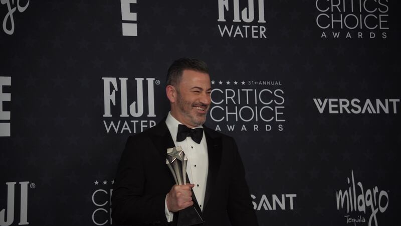 Jimmy Kimmel wears a tuxedo and holds up the award while the background says 31st Annual Critics Choice Awards.