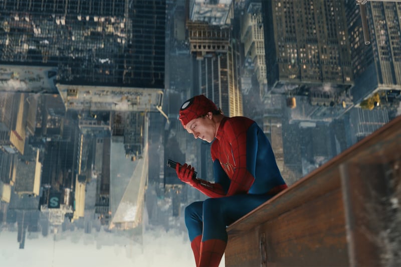 Tom Holland as Spider-Man is hanging upside down from a tall building in New York City dejectedly staring at his phone with his mask off and suit on.