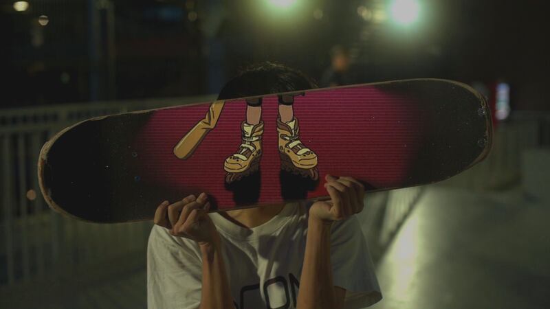Photo of a person holding a skateboard in front of their face. The skateboard shows someone's feet wearing rollerblades.
