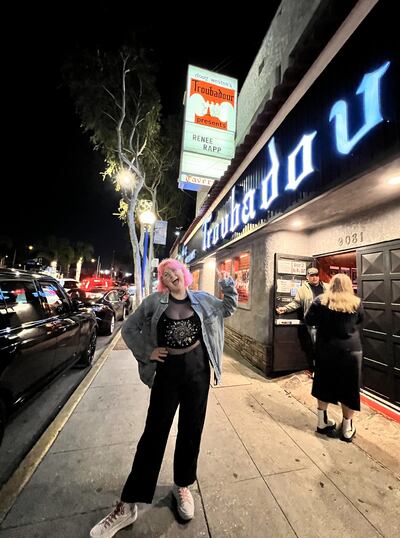A photo of Miga standing in front of the marquee at the Troubadour theater. She is pointing up at the sign.
