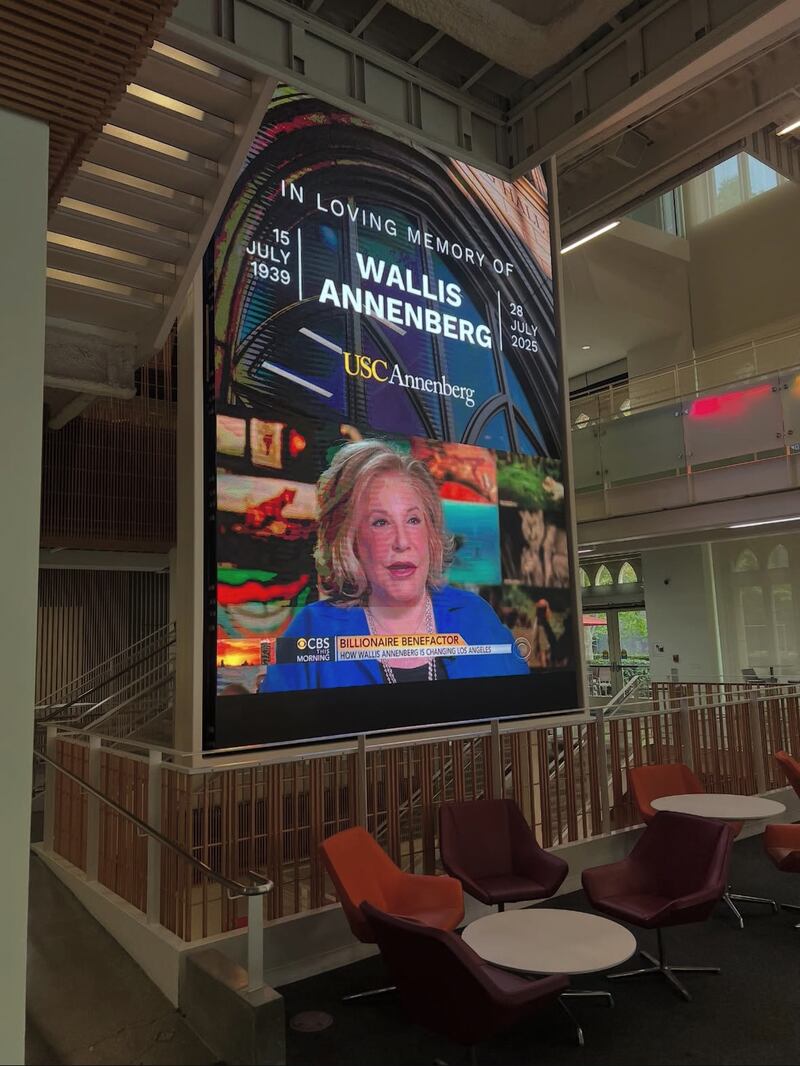 A huge screen in the center of a university building features a graphic with an image of Wallis Annenberg speaking in a CBS interview with the words "IN LOVING MEMORY of WALLIS ANNENBERG, 15 July 1939 to 28 July 2025."