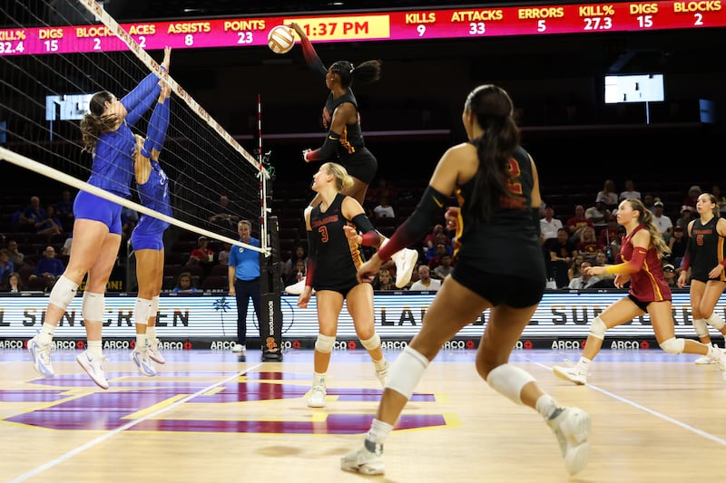 Rylie McGinest rises up to spike a volleyball over a net while teammates watch on. Two Creighton volleyball players jump and attempt to block the shot from the other side of the net.