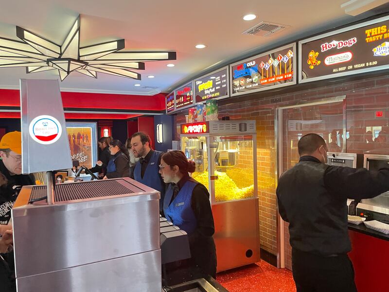 Photo of a counter with people in blue vests behind it next to a large popcorn machine and menu