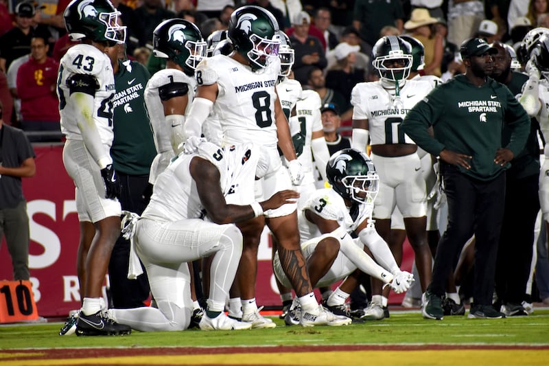 Michigan State platers kneel on their sideline wearing white and green football uniforms.