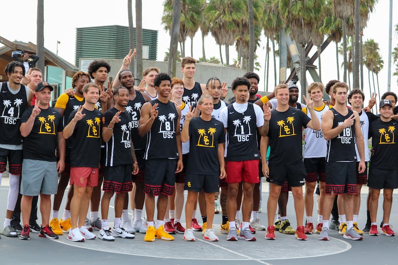 USC men's basketball (players, coaches and support staff) poses for a photo on the main Venice Beach basketball court. They wear black and white USC practice jerseys and several give the 'V for Victory' symbol with their hands.