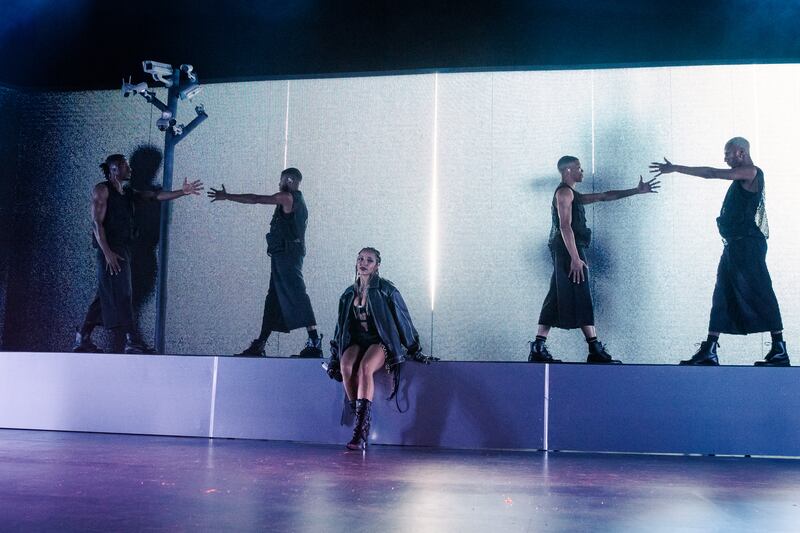 Photo of a woman singing in stage next to four dancers in front of a white screen