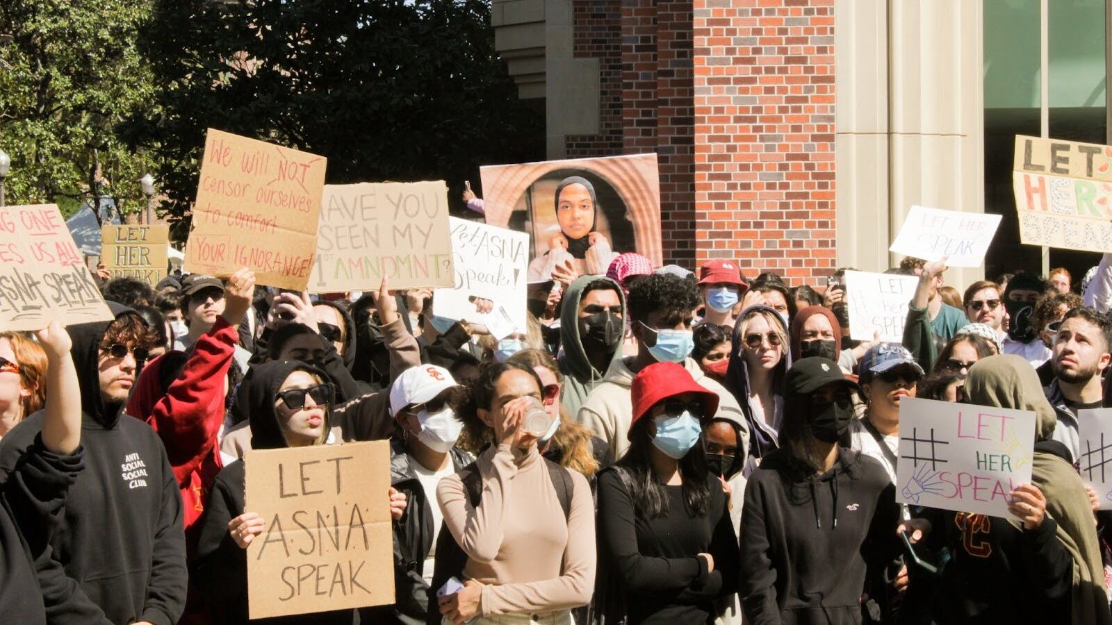 Hundreds of masked protestors in black wearing various masks, glasses and face coverings gather in front of a large brick building and some trees holding up cardboard signs.