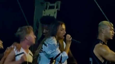 Tate is wearing a bedazzled jersey and singing into a microphone. There are two male dancers in jerseys near her.