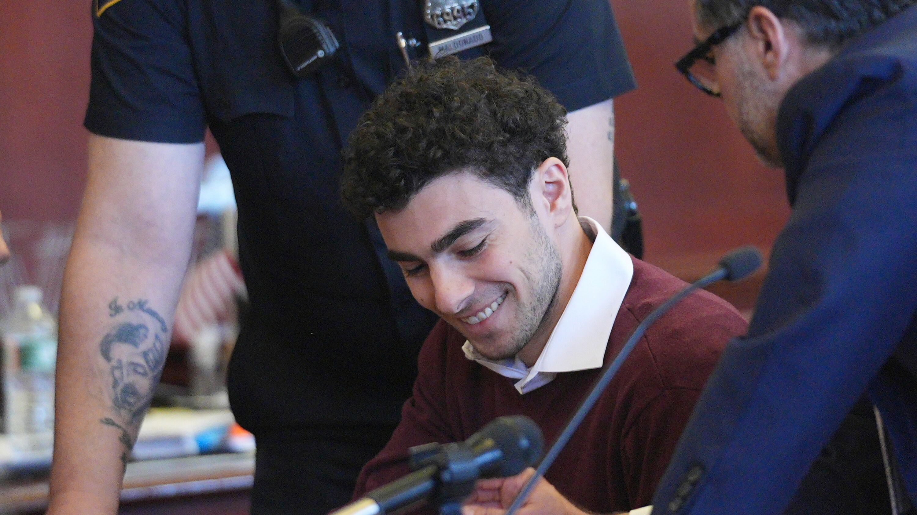 A man in a red sweater in a courtroom smiles while looking down, a cop over his shoulder.