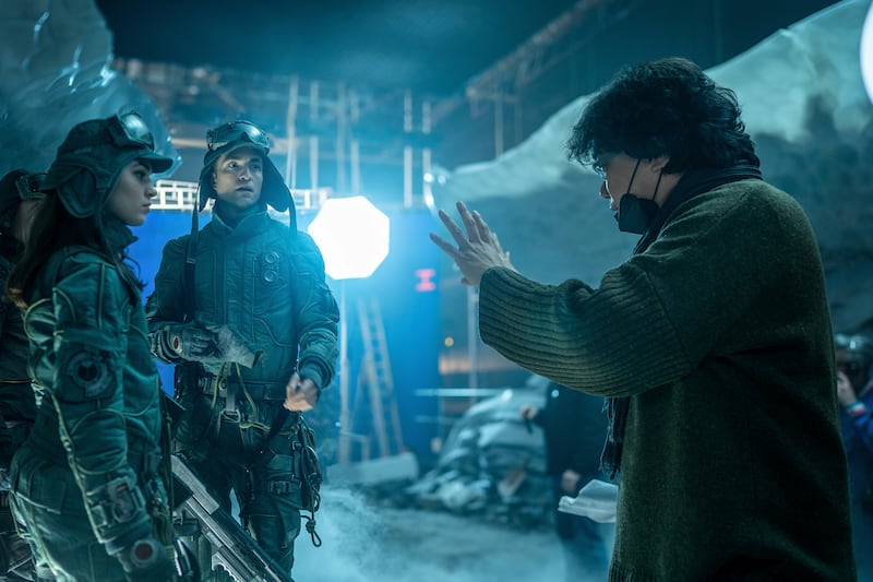 Photo of a woman and man in a costume with goggles next to a man wearing a sweater and mask directing them