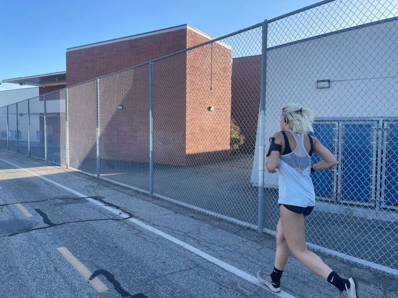Wearing a white tank top and blank running shorts, a woman with ice blonde hair pulled back runs on the sidewalk next to a large fence.