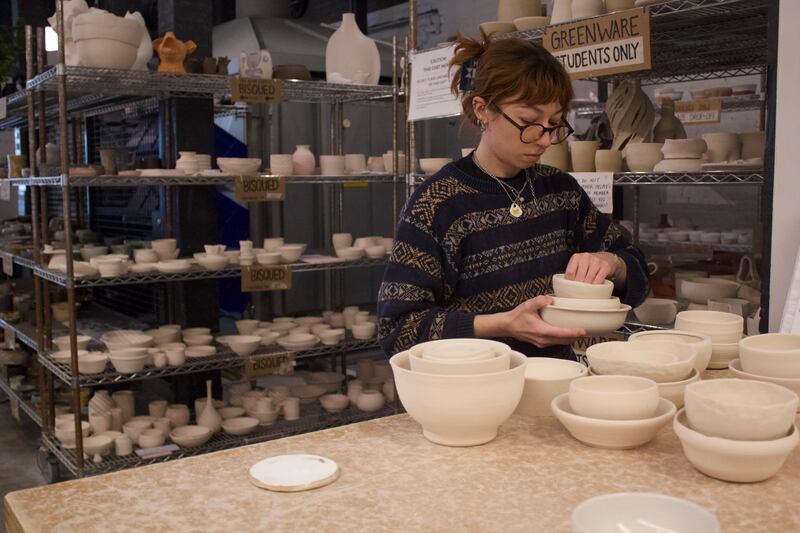 Photo of Studio Manager Bailey Walters organizes unglazed pottery. Shelves of student-made greenware sit in the background. (Photo by Ko Mosley)