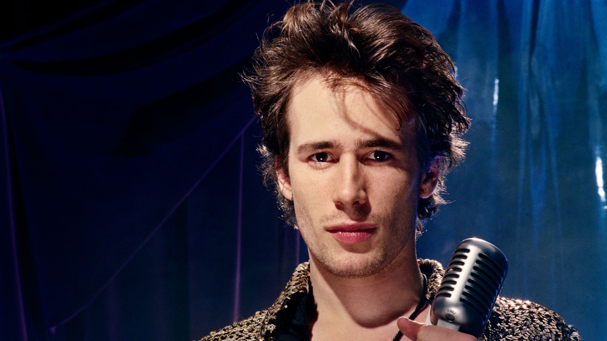 A young Jeff Buckley eyes the camera, with a microphone in his hands. His brown hair is tussled.