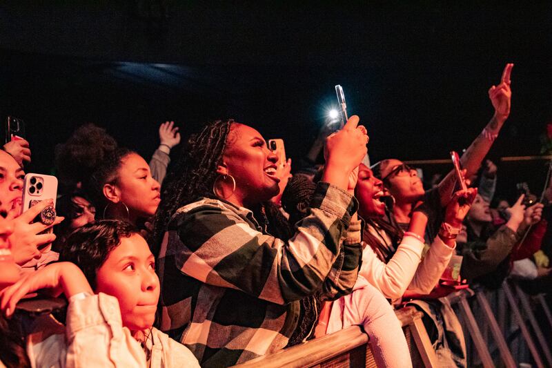 A photo of fans in the audience at the concert smiling and recording videos and taking photos of the performer on stage.