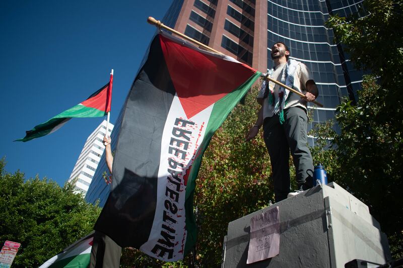 A protester stands above the crowd in front of the Israeli Consulate holding a flag saying "Free Palestine".
