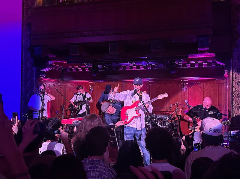 Rex Orange County wears jeans, an unbuttoned shirt and a hat. He is speaking into a microphone while pointing at the crowd. He has a red electric guitar around his chest.