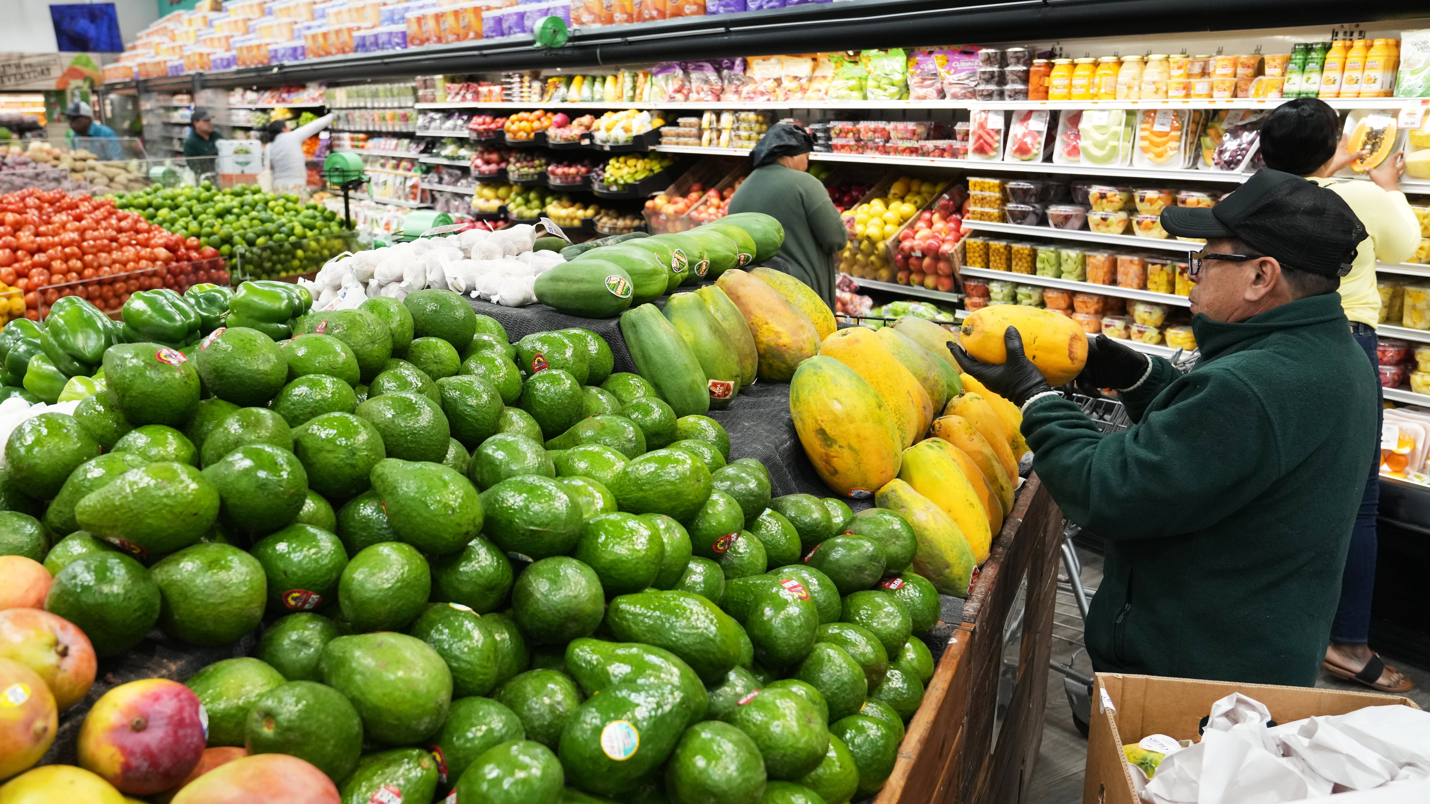 Photo of a man stocking papayas in a supermarket, surrounded by limes and peaches. Food can be seen behind him on shelves.