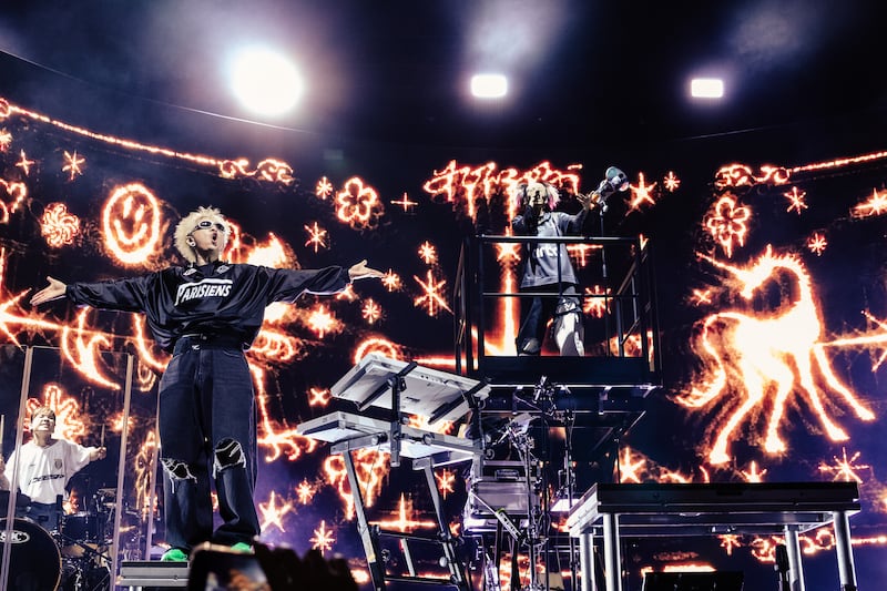 Photo of a band standing on a stage in front of a screen showing golden lights shaped like stars, smiley faces, and unicorns