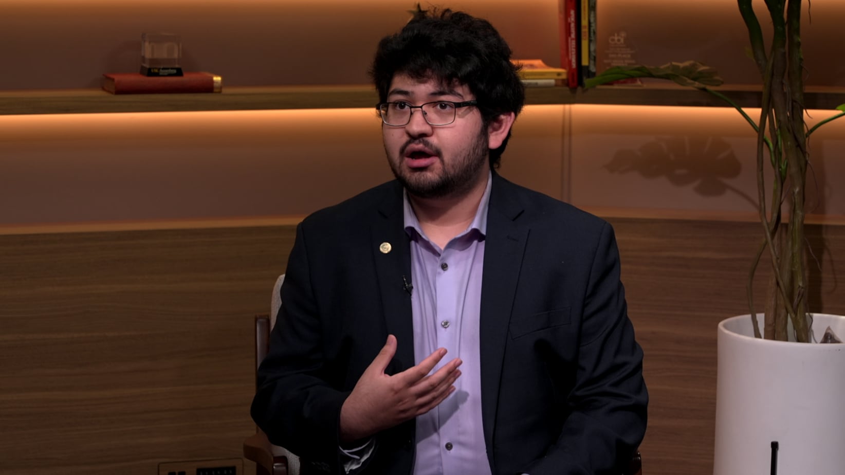 Moy is talking while wearing a black suit jacket, a light blue undershirt and black glasses.