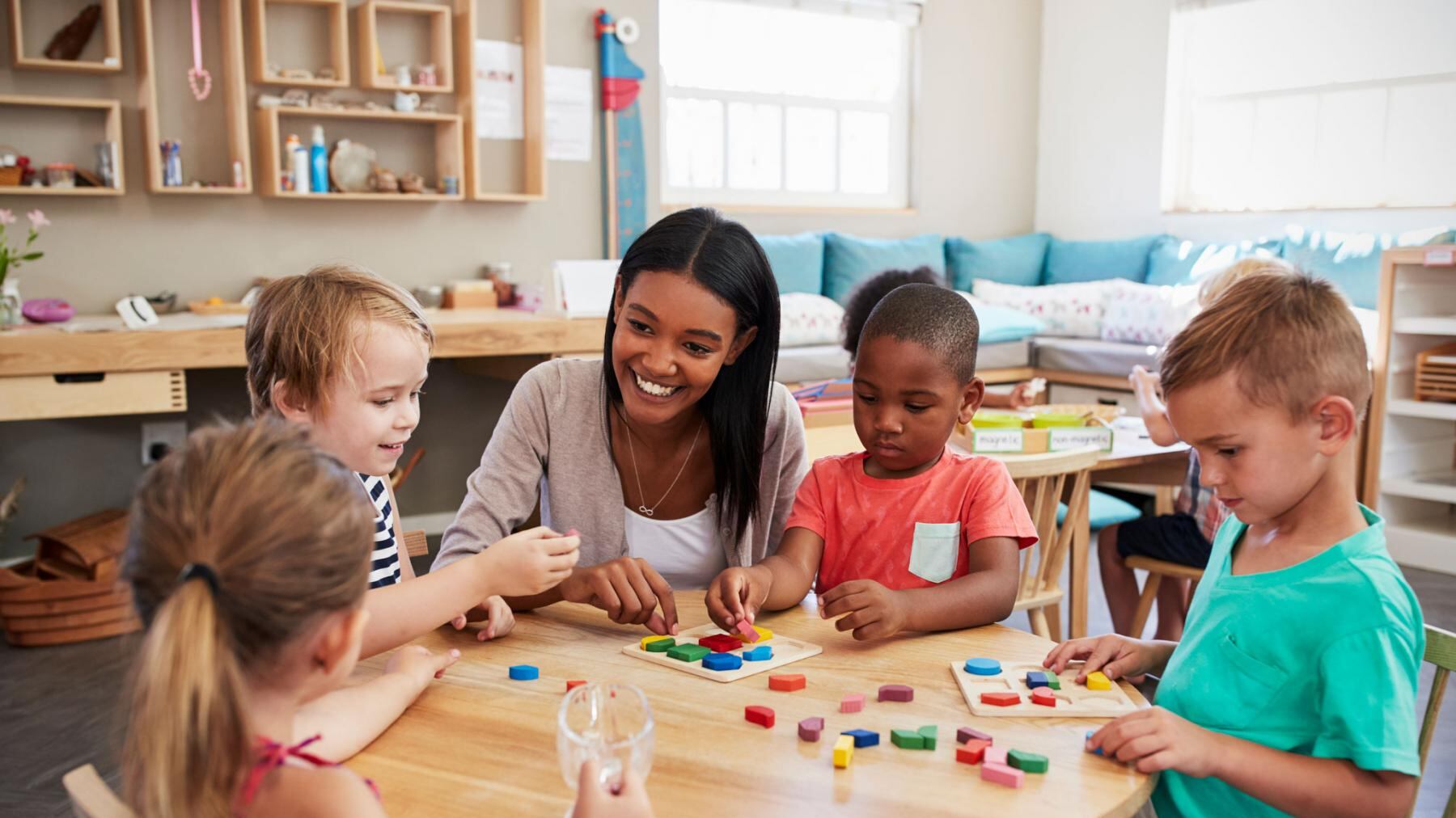 A photo of a day care room full of children sitting around a table alongside their caregiver.