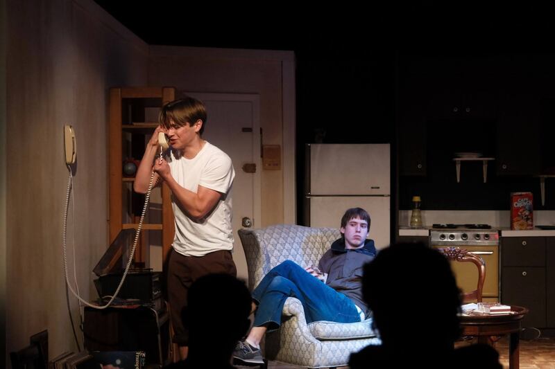 Onstage, one person in a white t-shirt holding the phone as if to call someone. One person is sitting down on a large chair behind him.