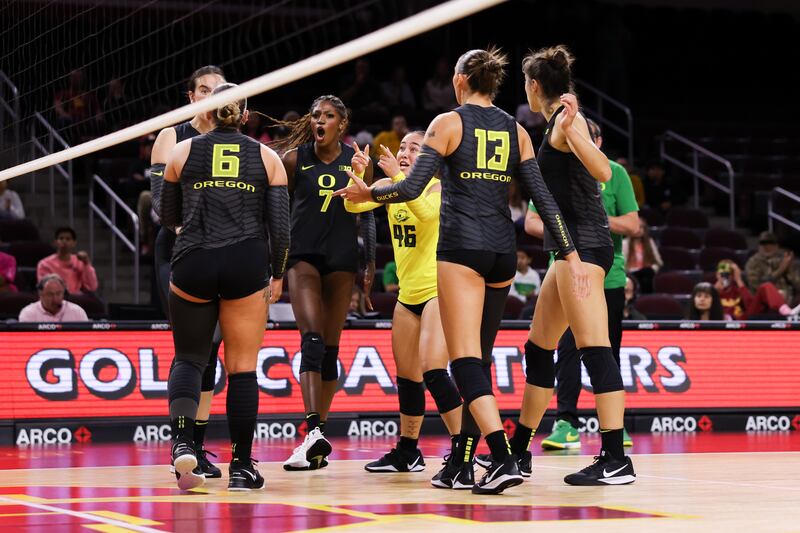 Oregon women's volleyball players celebrate in a huddle after a point. They wear black volleyball uniforms with yellow and green accents.