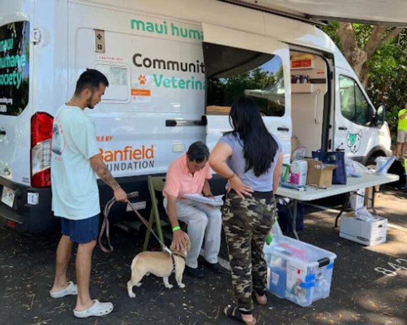 Photo of three people with a dog in front of a white van and a table