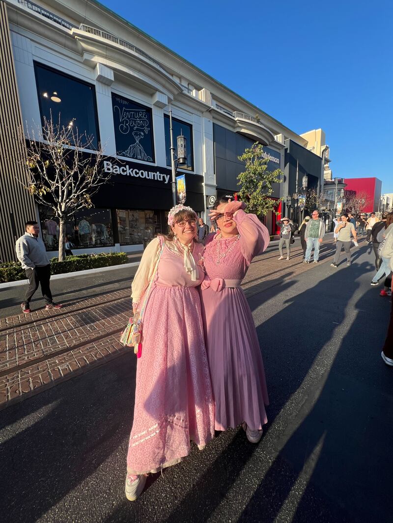 Two women smile while standing next to one another wearing long pink dresses. The details of the dress appear historic.