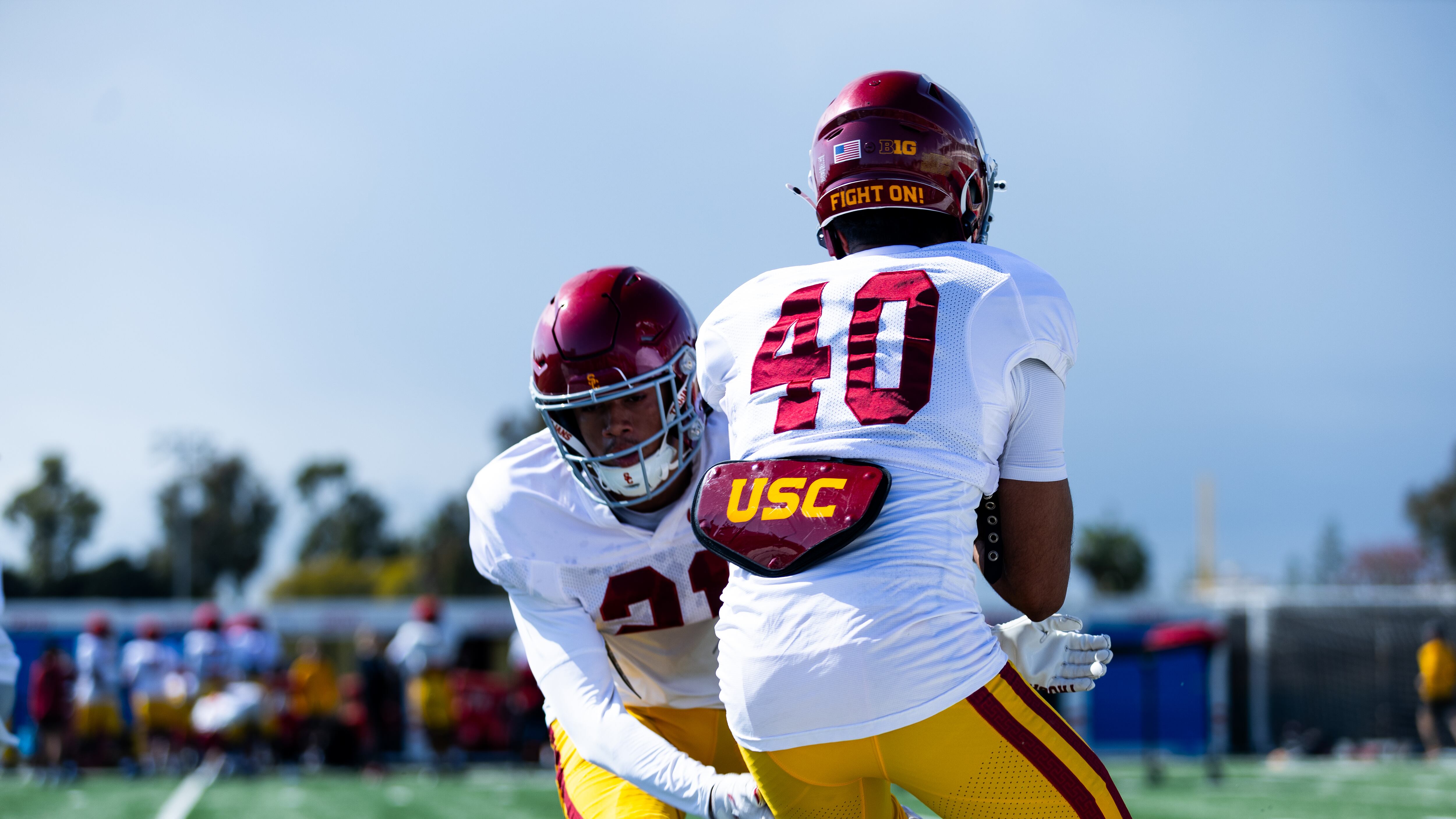 Two USC football players stand in the foreground as if about to tackle each other; one faces the camera while the other (40) faces away. Both are wearing white football jerseys with cardinal helmets and gold pants.