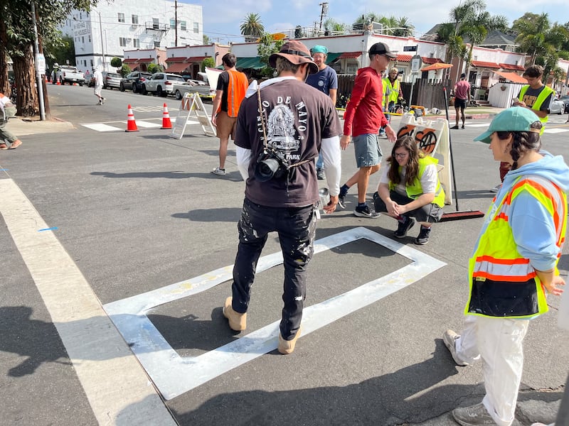 Volunteers in bright-colored vests place down a stencil to paint part of a crosswalk.