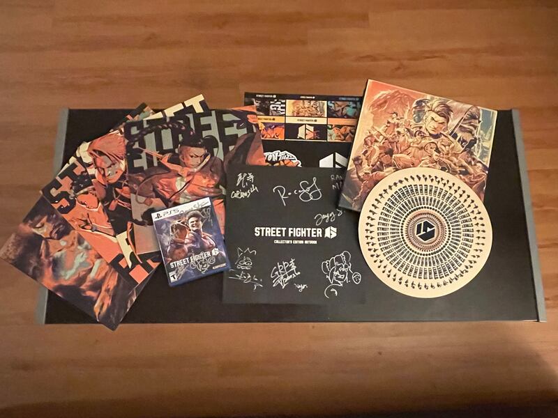 Photo of artwork on a table next to a PS5 copy of "Street Fighter 6" and a black book with signatures on it