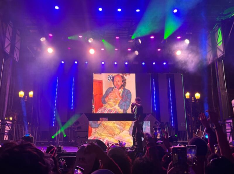 There is a photo of Sikowitz on the stage's digital screen. Matt is on stage talking to the audience. The lights are blue and green.