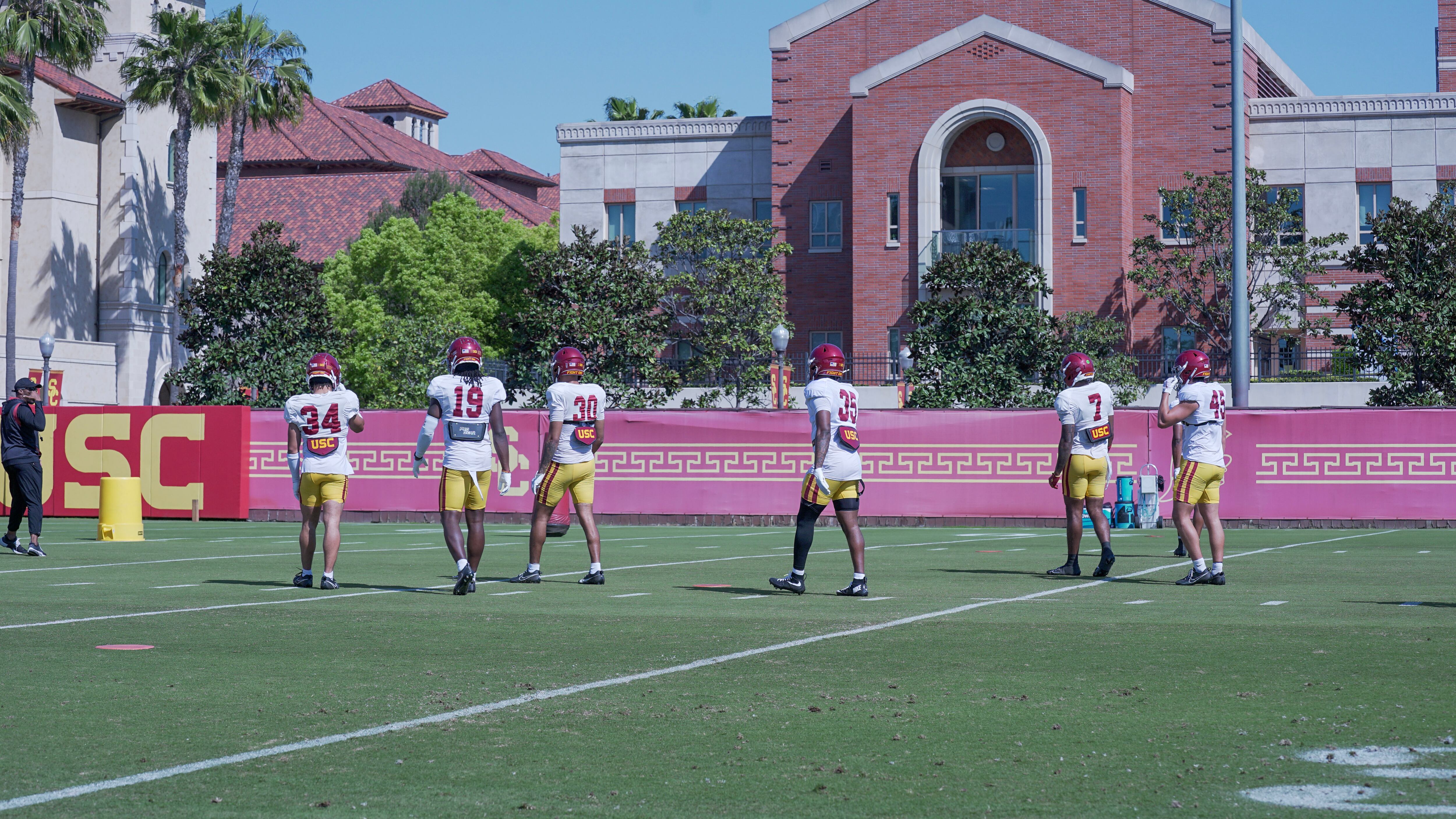 A photo of six USC players, all wearing white jerseys, with their backs to the camera getting ready for a drill.