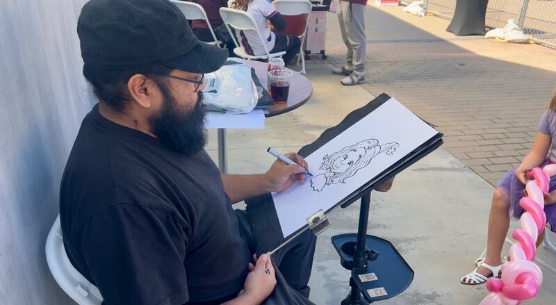 Julio is wearing a black shirt and a black hat. He sits in a chair with an easel in front of him where he sketches a caricature of a young girl sitting in front of him.