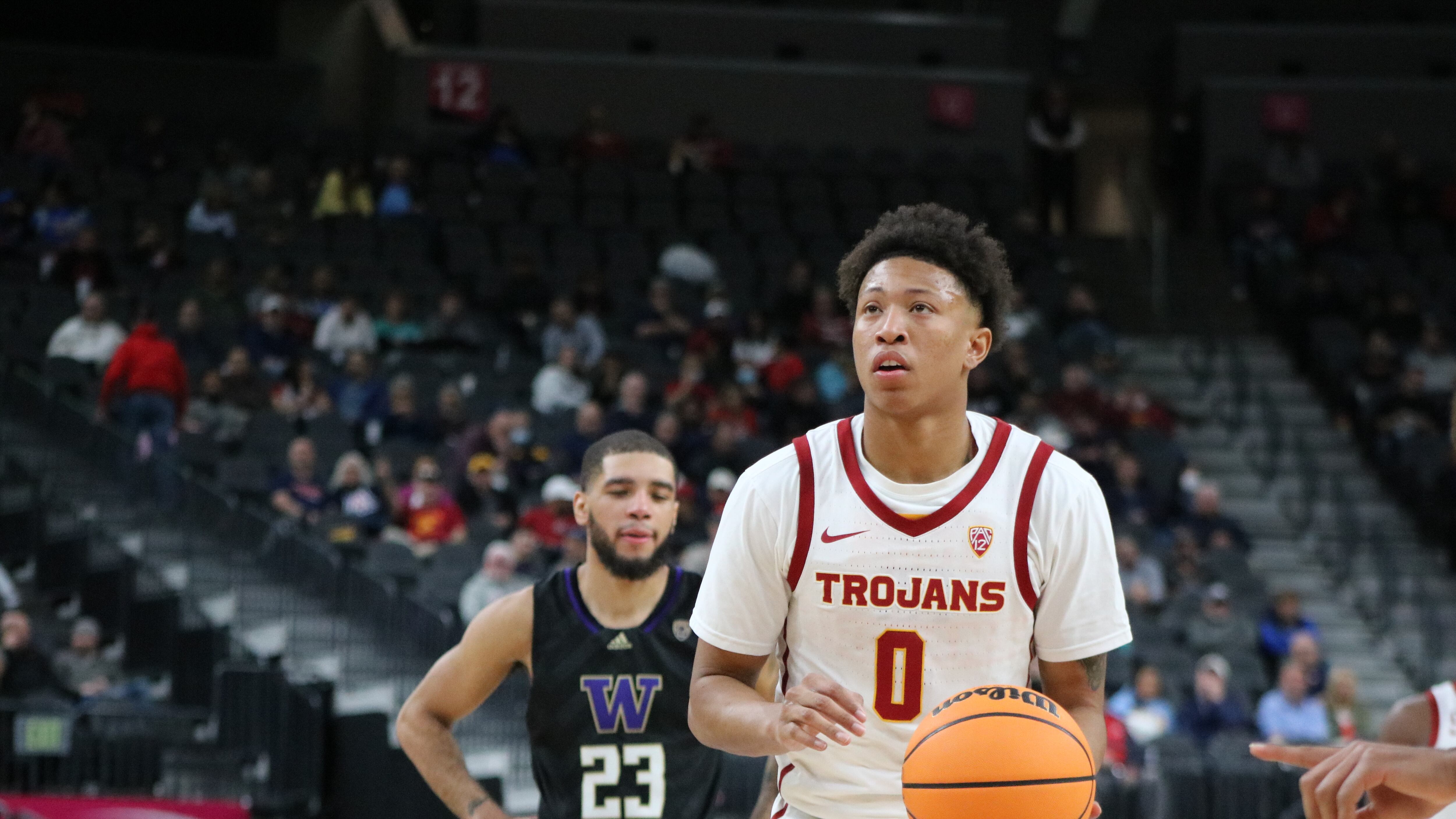 A photo of junior guard Boogie Ellis going through his free-throw routine. He's wearing a white USC jersey with the word "Trojans" in Cardinal lettering, and holding the basketball in his left hand while looking at the rim.