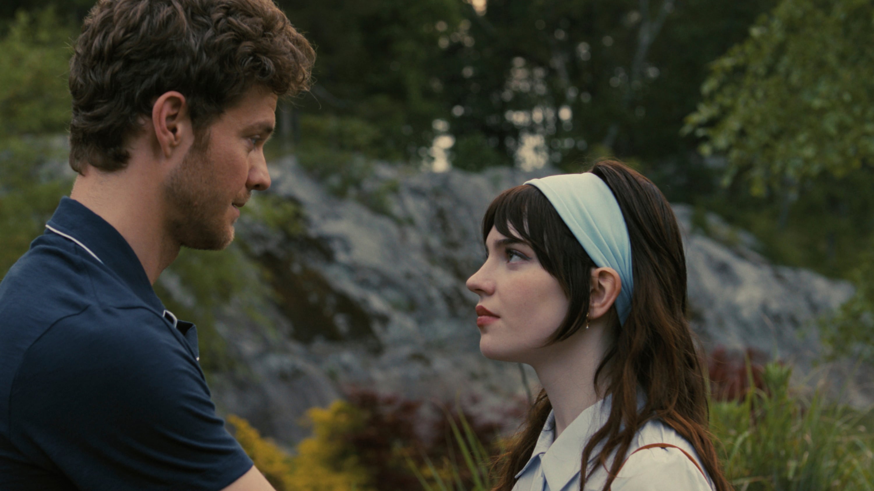 Quaid is wearing a navy blue polo while looking in the eyes of Thatcher. Thatcher is wearing a light blue headband. They are in a forest