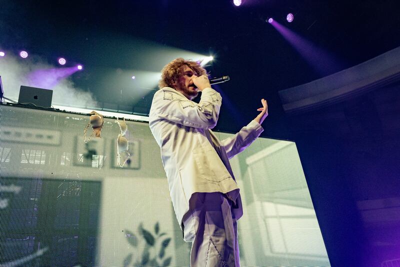 A photo of Yung Gravy singing into a microphone on stage. On the stage in the background, there are bras hanging on the set.
