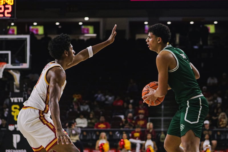 Jerry Easter II (8) puts a hand in the face of a Cal Poly player holding the basketball. He wears a white basketball uniform with cardinal and gold accents while the Cal Poly player wears a green basketball uniform with white accents.