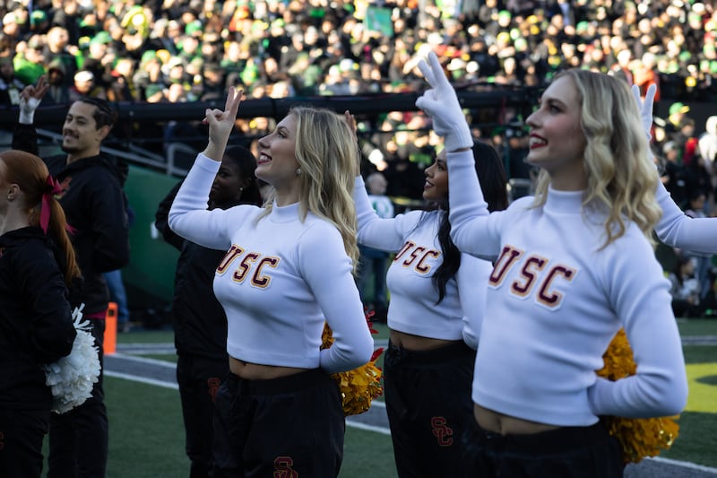 USC cheerleaders smile and hold up a 'V for victory' symbol. They wear white long-sleeved shirts with 'USC' on the front in cardinal and gold.