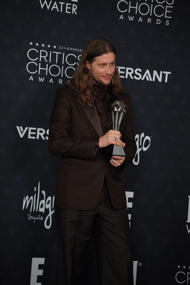 A composer in a brown suit smiles with an award in the press room.