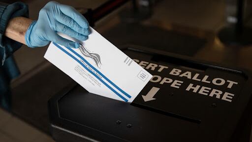 A photo of a hand wearing light blue latex gloves inserting a ballot into a drop box.