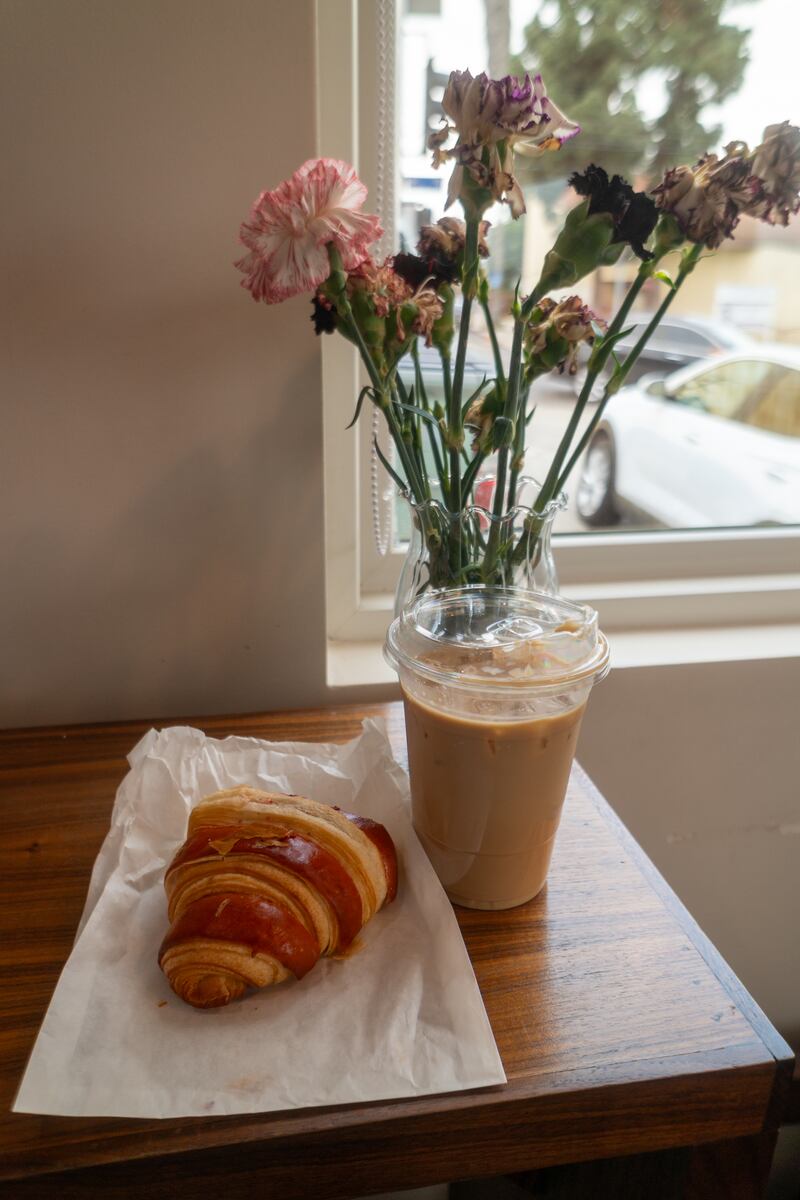 Photo of a croissant and a latte with flowers in a vase in the background.