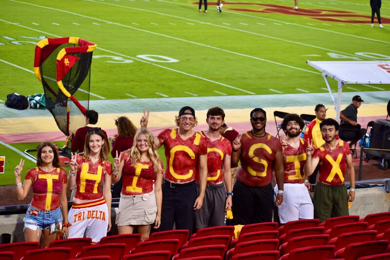 Eight USC students stand in the student section with the field in the background. They are all painted red with yellow letters; each is painted with one of the letters in the term 'T-H-I-C-C-S-I-X'.