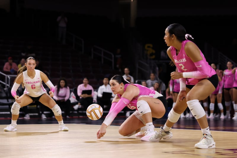 Dani Thomas-Nathan (15) crouches down low on one knee to dig a volleyball and prevent it from hitting the ground. She is surrounded by Trojan teammates on both sides.