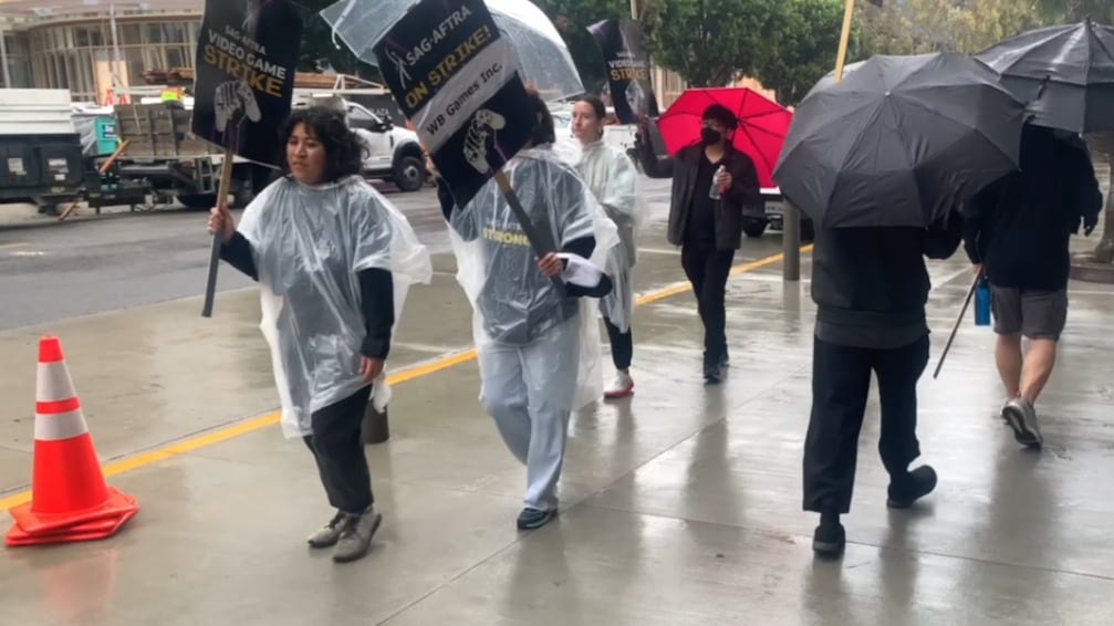 Protestors march on a sidewalk with signs in their hands while it rains.