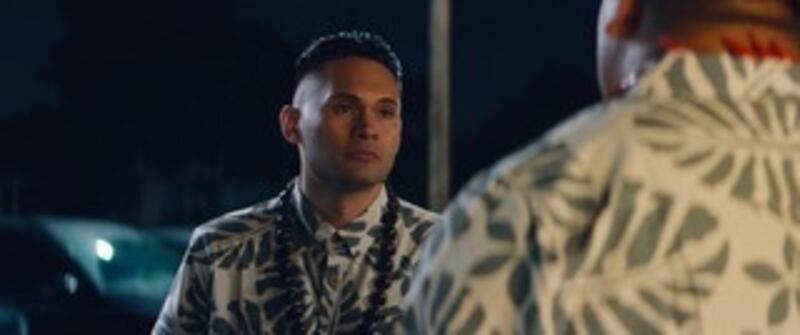 Film still of a man wearing a button-up with a leaf pattern and a necklace with large black beads on it