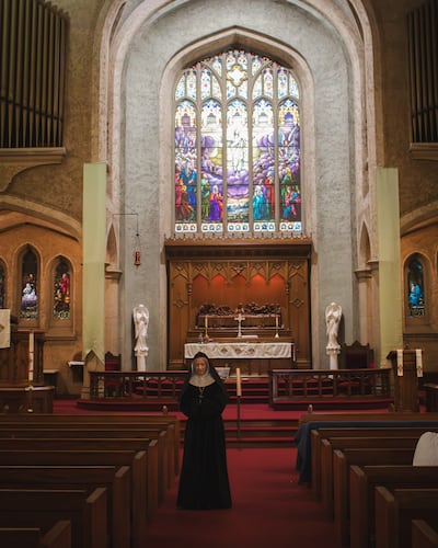 A nun actress stands looking at the ground with a church behind her.