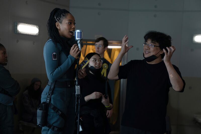 Photo of a woman with a gray costume and a microphone next to a man with a black shirt and glasses