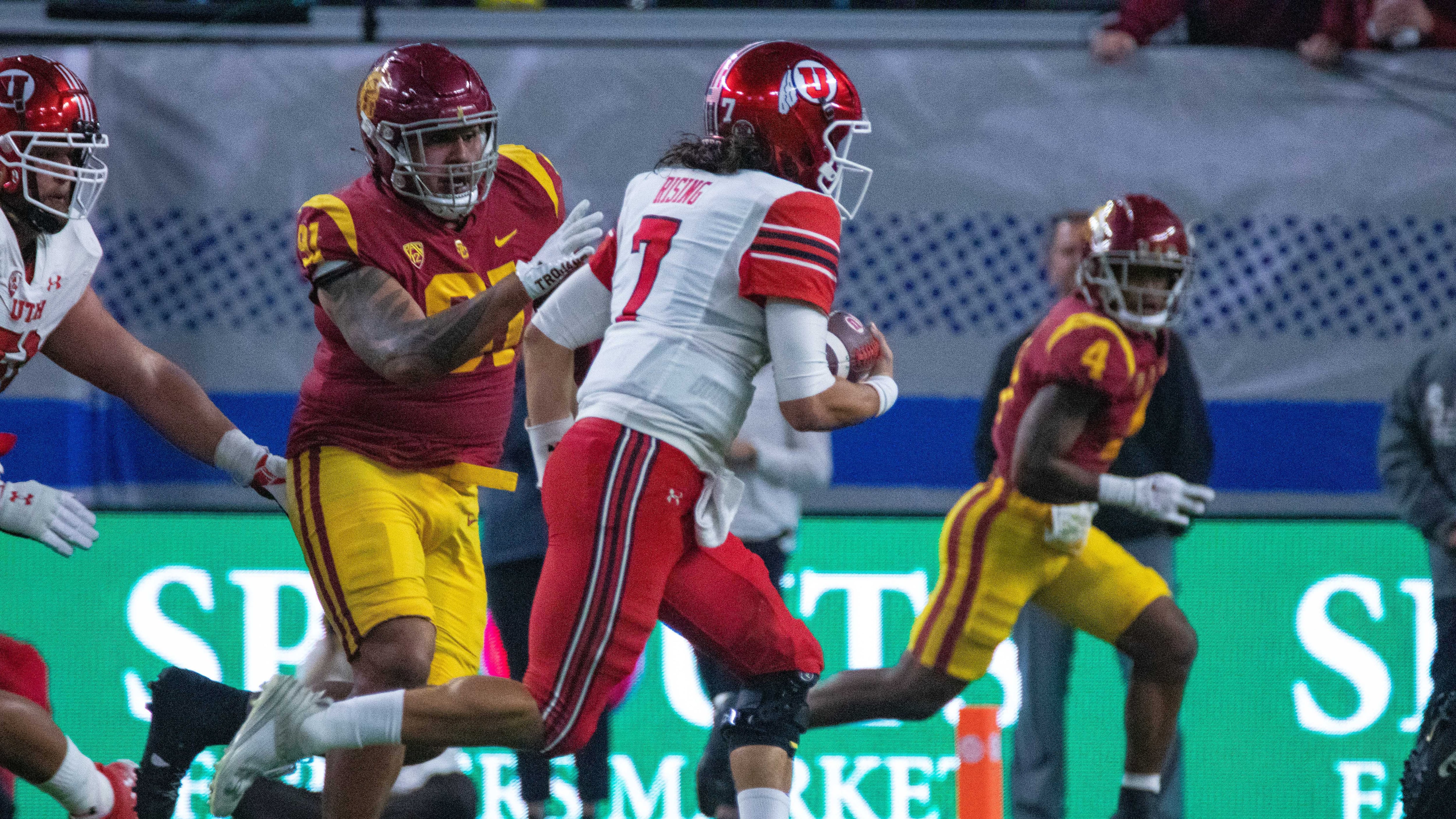 A Utah player in red pants and a white jersey, carrying the ball, running from the USC defensive player in a cardinal jersey and gold pants.